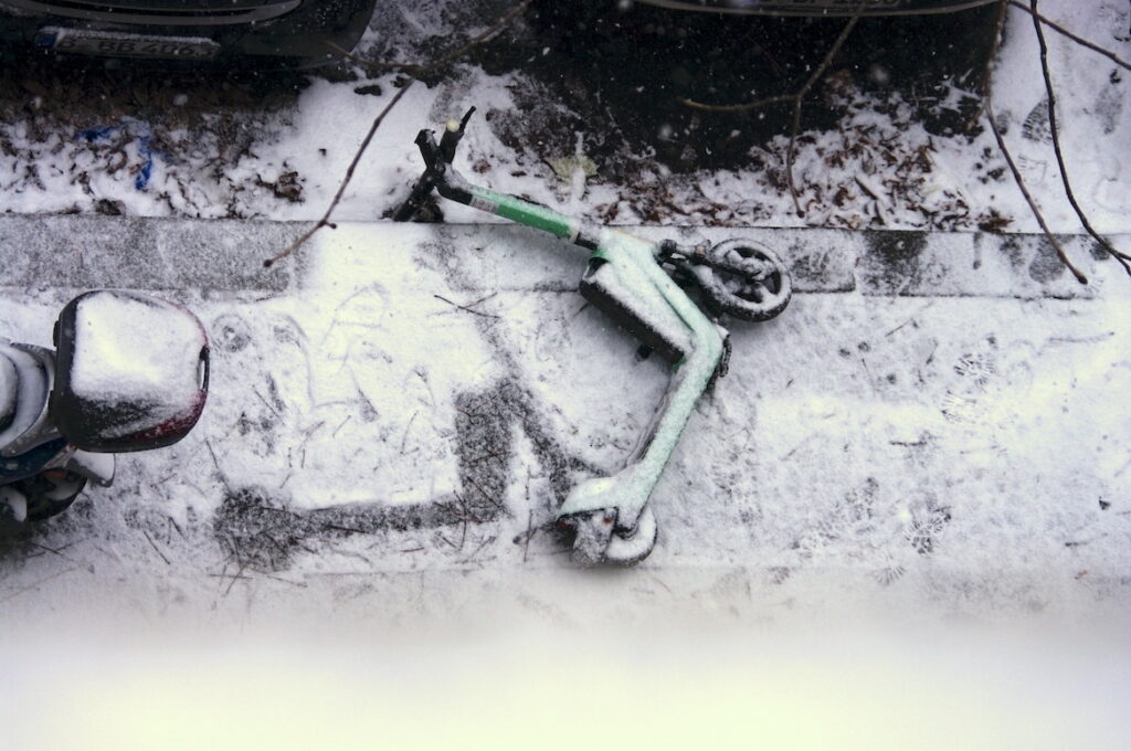 Ein E-Scooter liegt schräg am Strassenrand, verschoben von seinem vormaligen Platz, der seine hinterlassene Form negativ im Schnee abbildet.